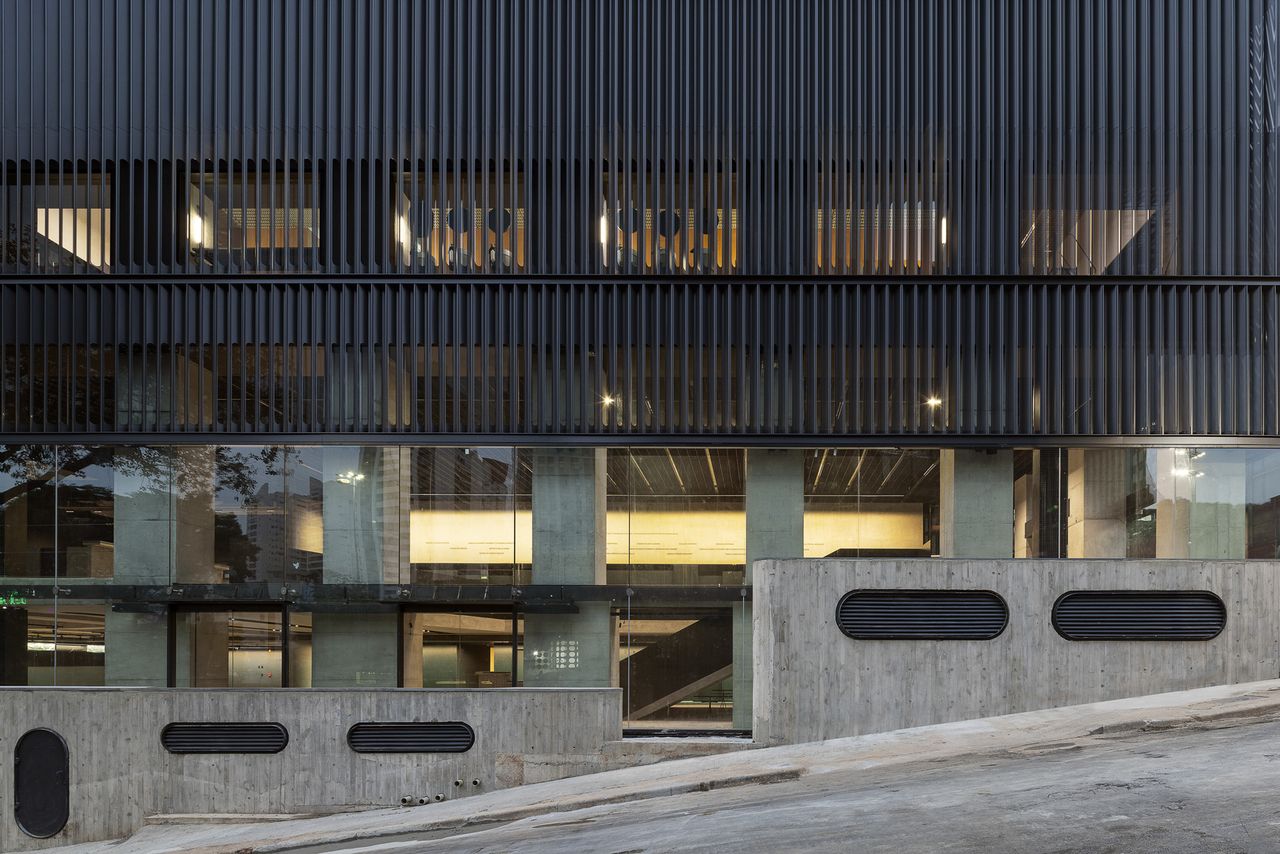 The warmly lit interiors of a brutalist building cast from cement, glass, and iron is captured at dusk from the side of a street.
