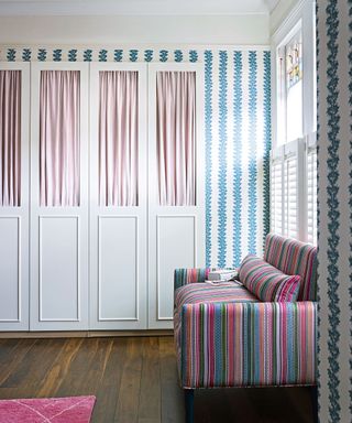 Bedroom with dark wooden floor colourful striped sofa, blue and white wallpaper and built in wardrobes, pink fabric curtains