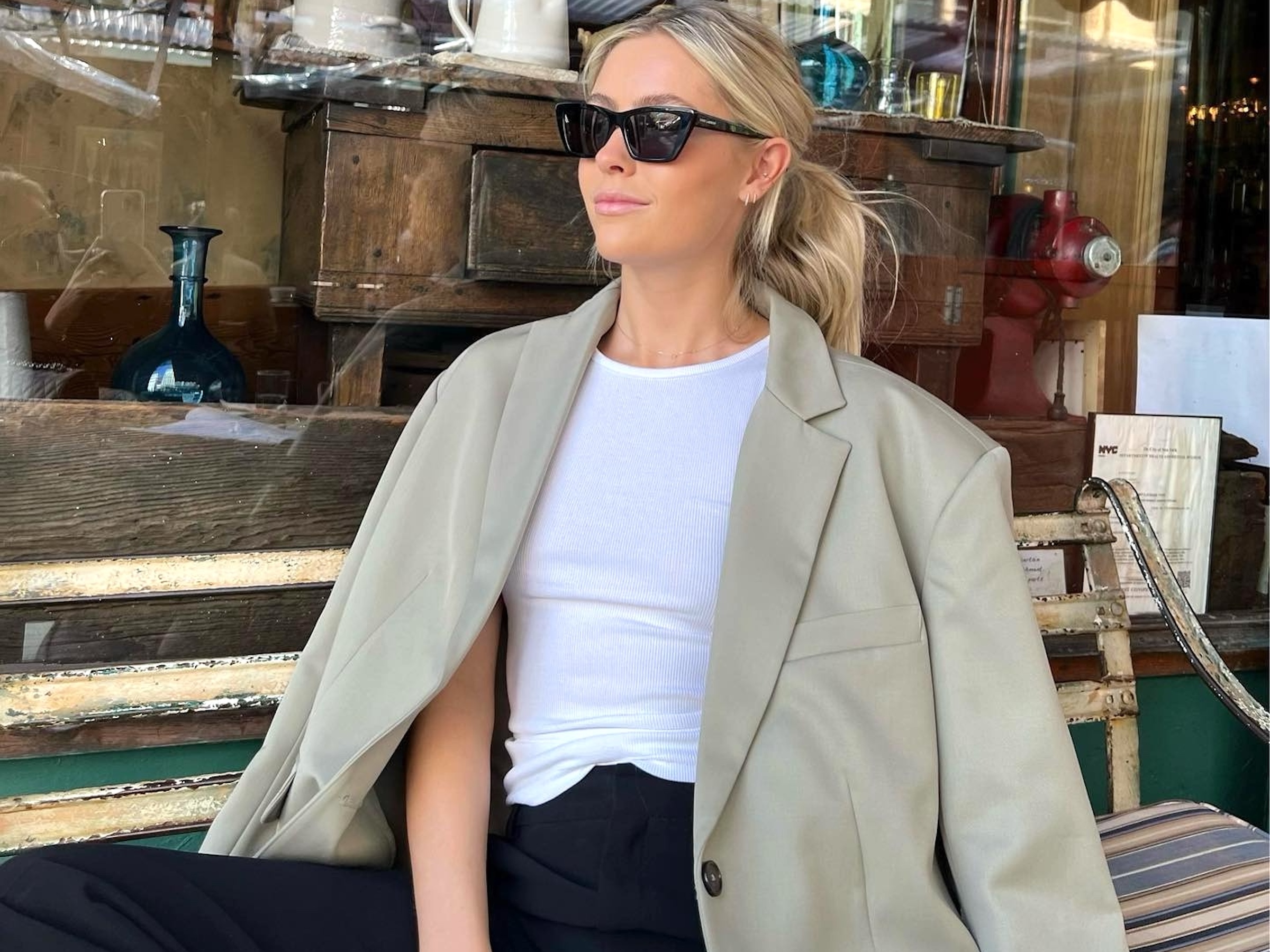 style influencer Fredrika Ekerot sits on a wooden bench outside a shop in NYC with a ponytail wearing black sunglasses, an oversize tan blazer, white tank top, and black pants