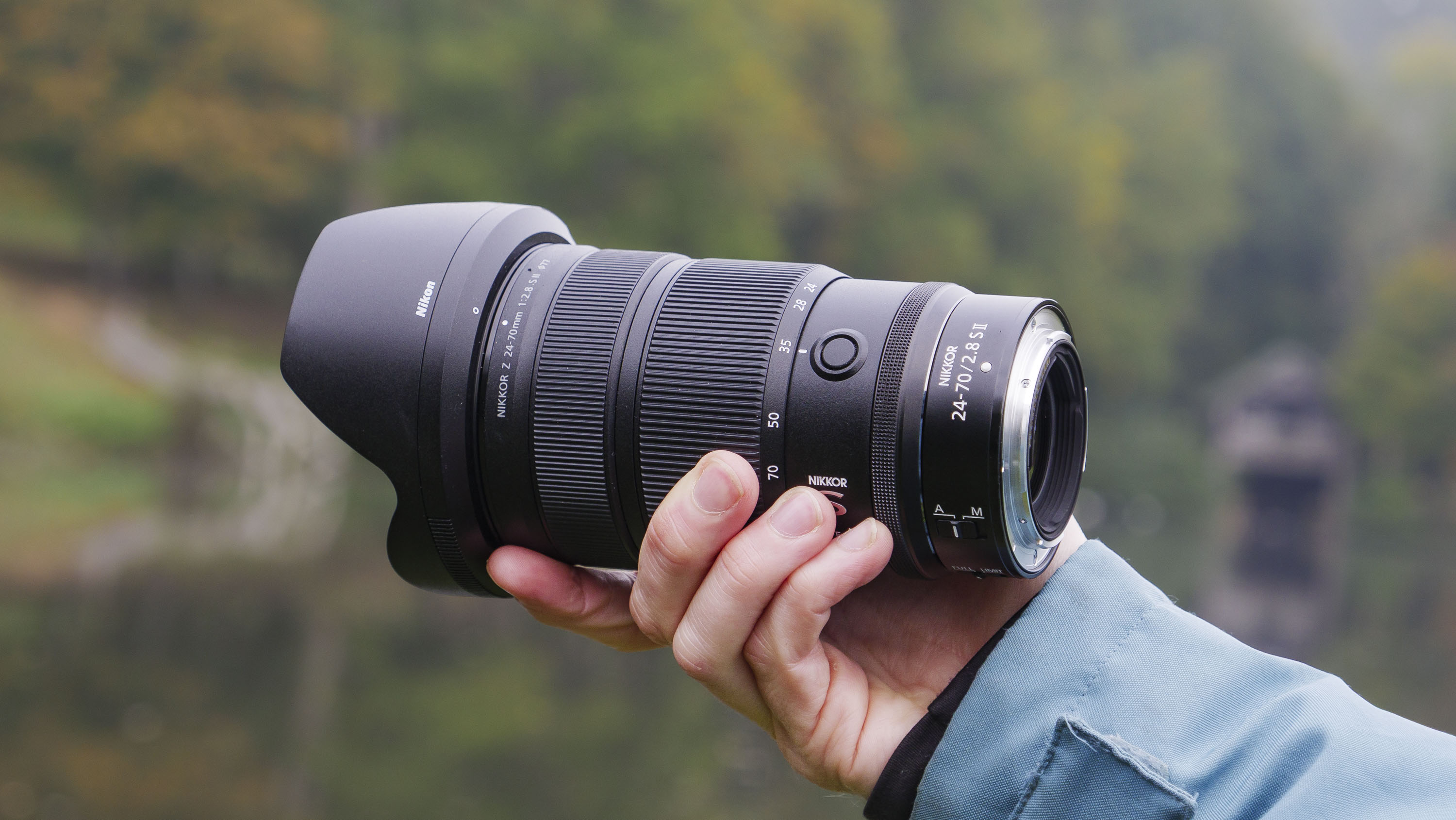 Nikon Z 24-70mm f/2.8 S II lens in user's hand, with autumnal lake background