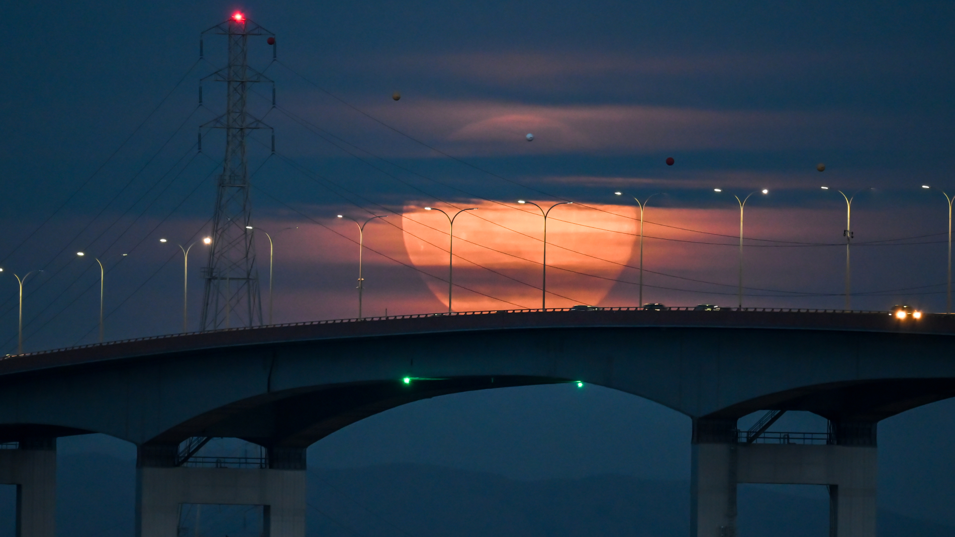 An orange full moon is pictured rising behind a bridge as clouds obscure the upper region of its disk. Streetlamps line the bridge and an electrical tower is positioned to the left of the image, with its cables falling across the moon&#039;s disk.