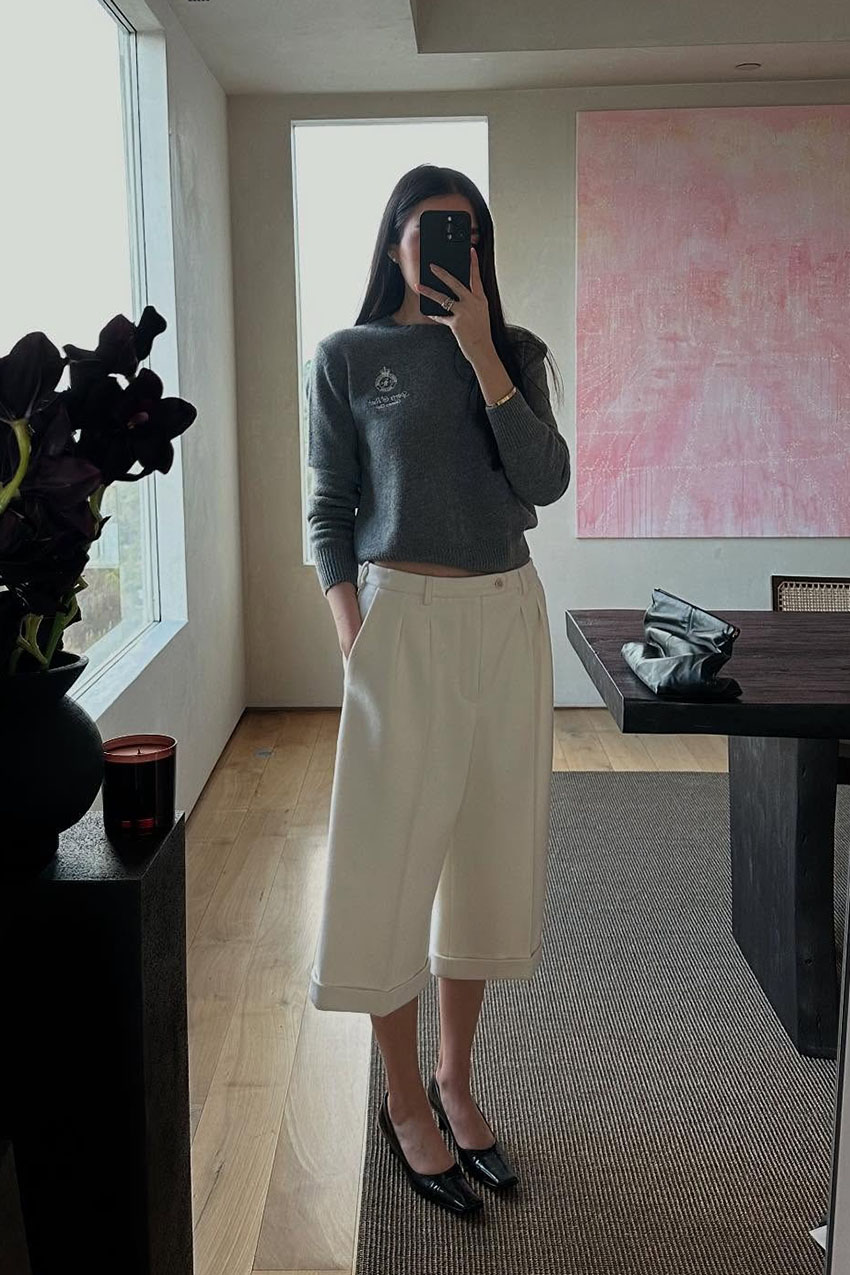A woman wearing a gray knit sweater, white culotte pants, and black pointed-toe flats