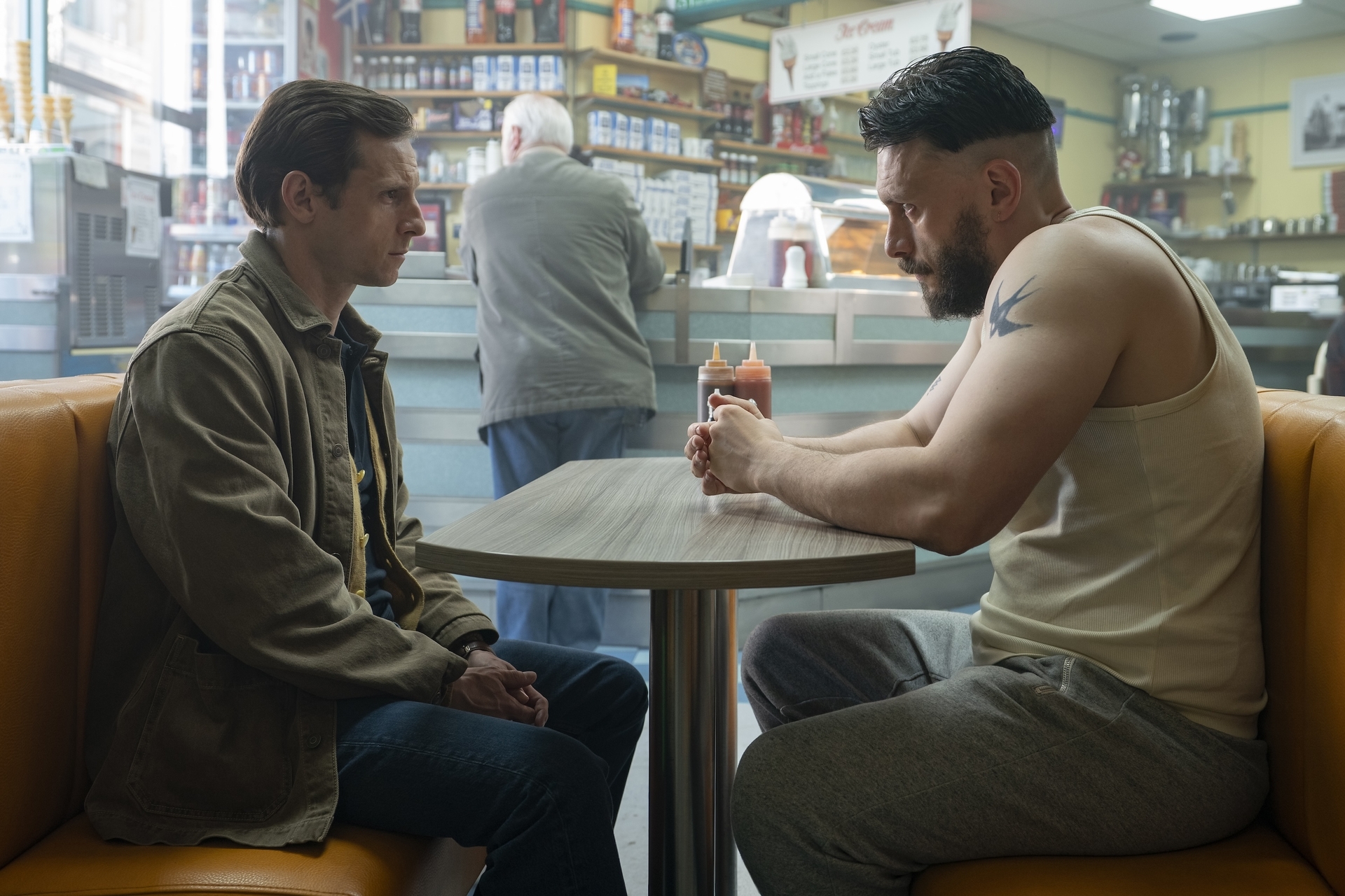 Two men (Jamie Bell and Richard Gadd) sit at a diner booth. A white-haired man stands at the counter behind them, looking at a back wall lined with shelves, in a first-look image for HBO's 'Half Man.'