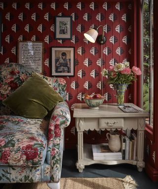 Lively living room with red patterned wallpaper, metallic wall lamp, large floral armchair, wooden side table with books and flowers, artwork on walls