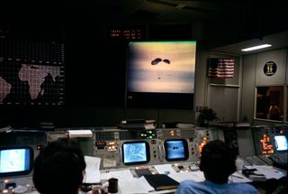 At NASA's Mission Operations Control Room in Houston, two flight controllers oversee consoles as they watch the Apollo 13 command module drop through the sky toward the south Pacific Ocean with three NASA astronauts on board. Behind the camera, a large crowd of people gather to watch the splashdown and recovery operations from the control room.
