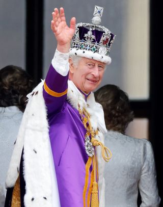 King Charles wearing his purple and white fur coronation robe and a crown waving