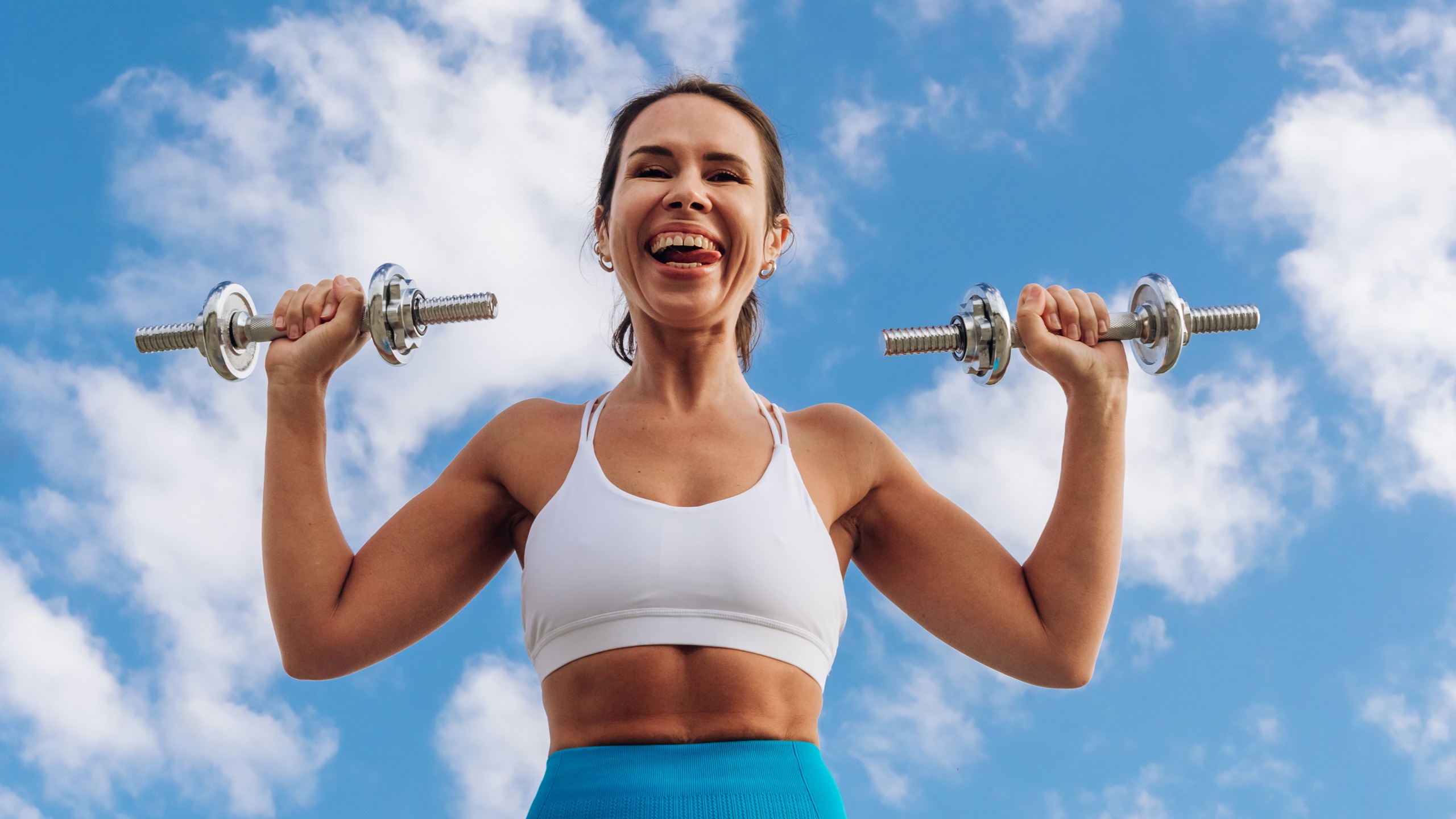 A woman holding dumbbells stands smiling, with her tongue poking out one side of her mouth. Behind her we see blue sky and white clouds.