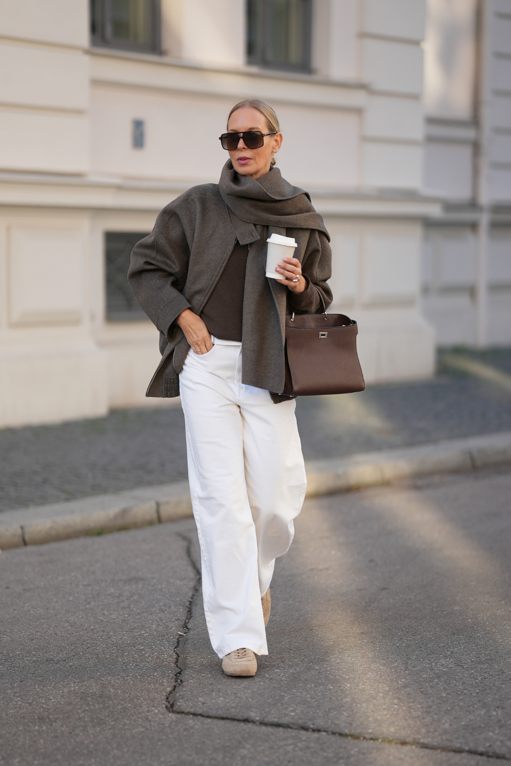 Mirja Klein is seen wearing an oversized grey brown wool bomber jacket with an elasticated waistband and built-in detachable scarf by Massimo Dutti; a brown wool sweater by Massimo Dutti; white wide-leg jeans with a straight fit by Massimo Dutti; beige suede trainers with a rounded toe by COS; a structured dark burgundy leather handbag with a top handle and silver hardware by Michino Paris; pilot frame sunglasses in black with a double bridge by Tom Ford; her blond hair is styled in a sleek low bun