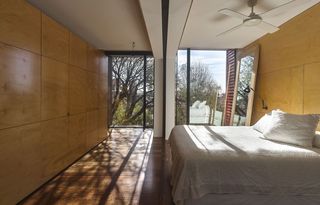 Alternative view of a bedroom at Moor Street House featuring wood panelled walls, wood flooring, a bed with white pillows and linen, a wall lamp, a white ceiling fan, a large mirror and floor-to-ceiling windows offering a view of the trees and blue sky outside