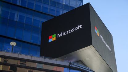 Microsoft logo and branding pictured on a sign on top of a New York City office building, with clear skies and skyscraper in background.