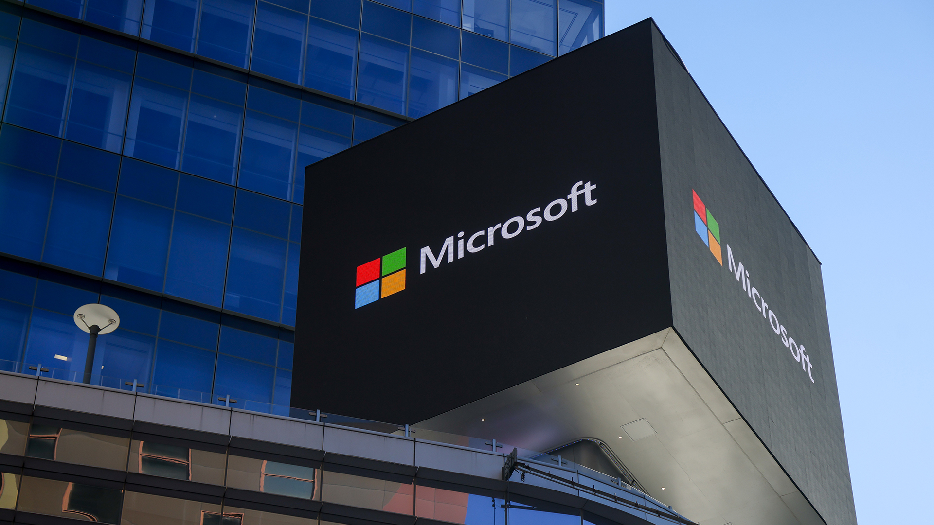 Microsoft logo and branding pictured on a sign on top of a New York City office building, with clear skies and skyscraper in background.