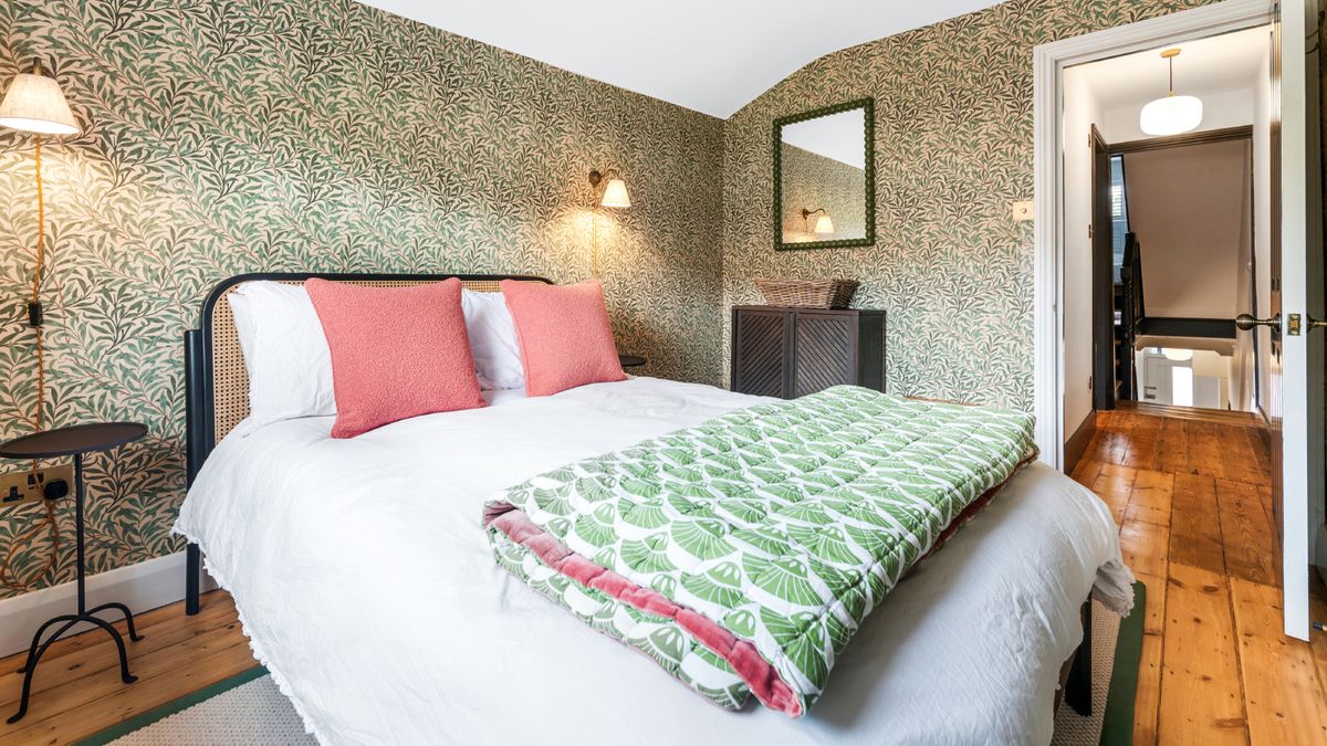 loft conversion bedroom with green floral wallpaper, wooden floor, black metal framed bed with white bedding, green and white throw with door open leading to small landing and stairwell