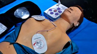 A medical dummy is shown lying on a blue sheet. Defibrillator pads are placed on the front and side of the chest and connected to a machine