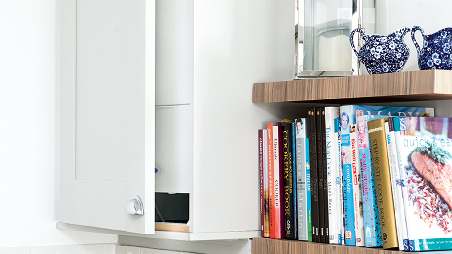 white kitchen with a cupboard open showing a boiler, beside open shelves with cookbooks