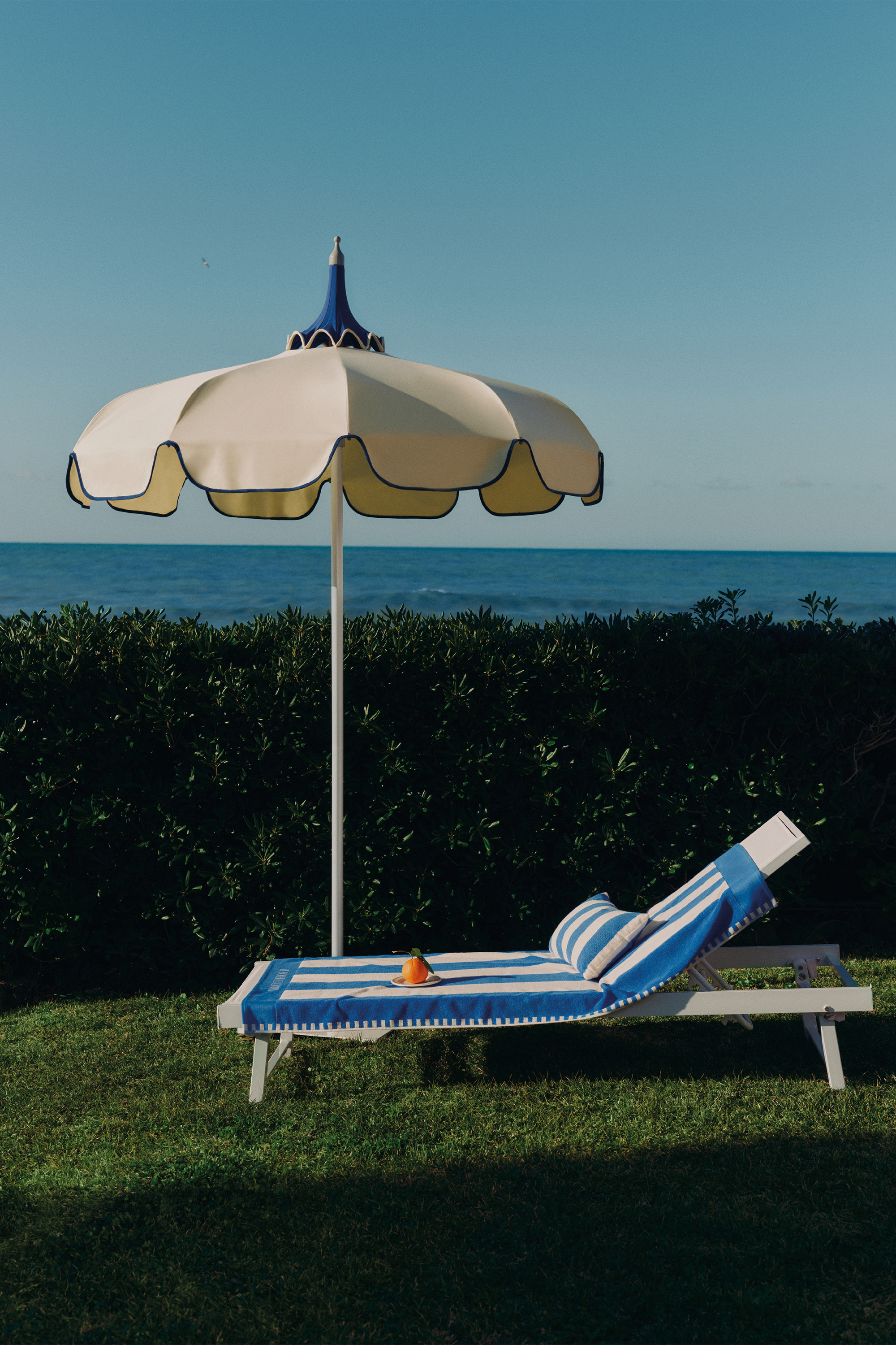 Image of a white lounger chair with a blue and white striped towel on it on a grassy lawn by the sea. There is also a white parasol.