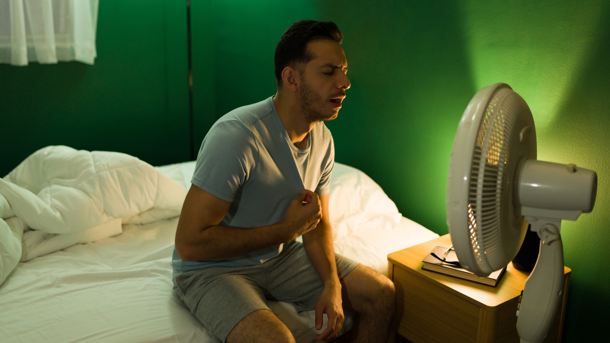 Sweaty man sitting on bed at night, facing an electric fan