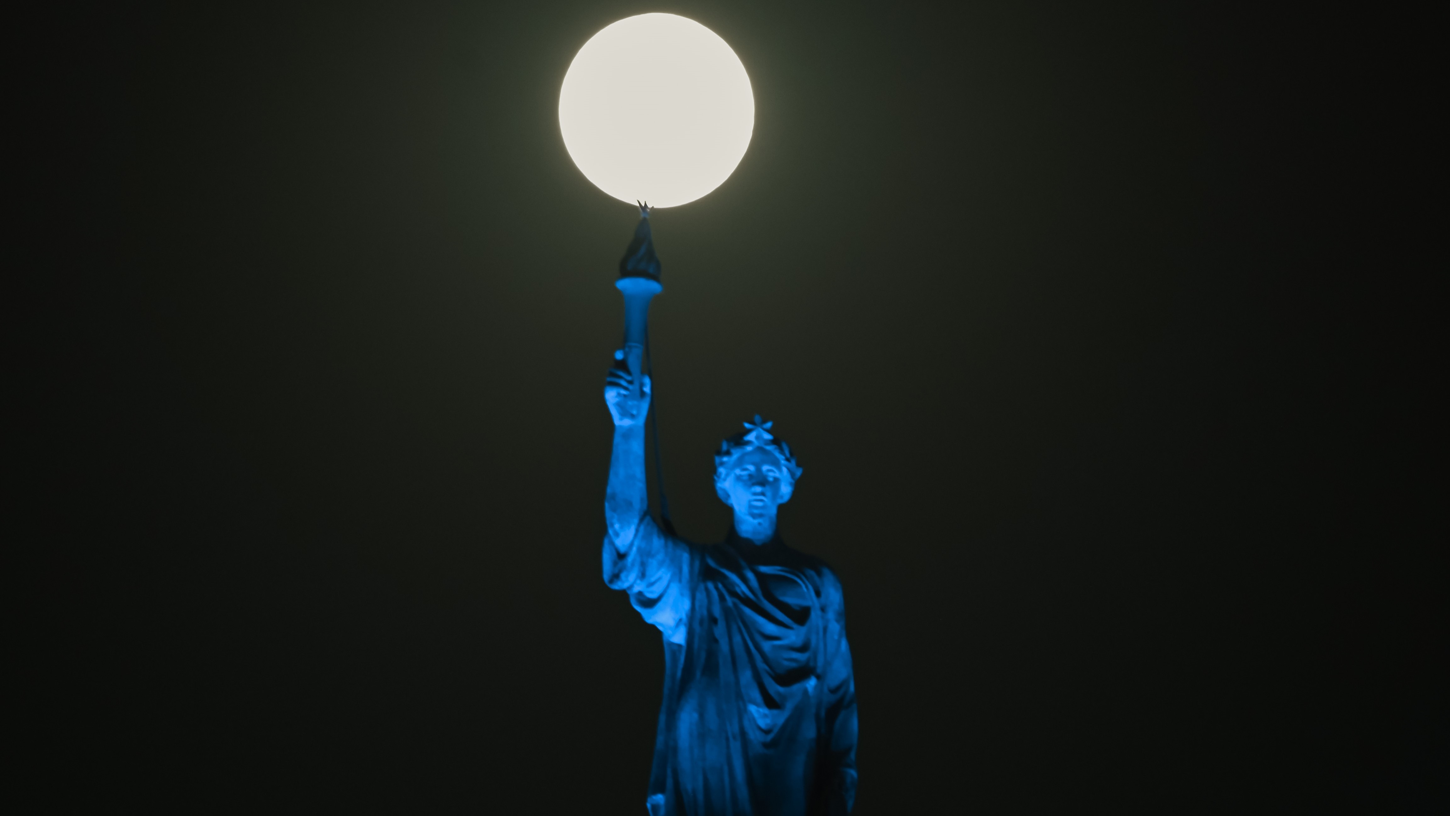 a statue holding up a torch is illuminated blue and atop of the torch the full moon shine bright.