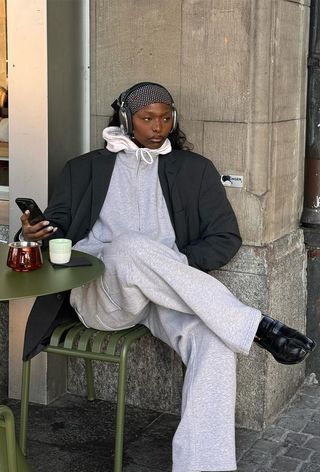 A woman sitting outside at a cafe with headphones wearing a white hoodie under a gray sweatshirt and gray blazer with matching sweatpants and Tabi loafers.