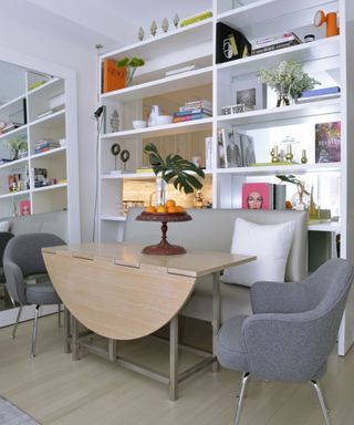 A dining room with a tall white bookshelf with plants and decor on it, a light wood drop leaf table with a red lamp with two gray tables either side