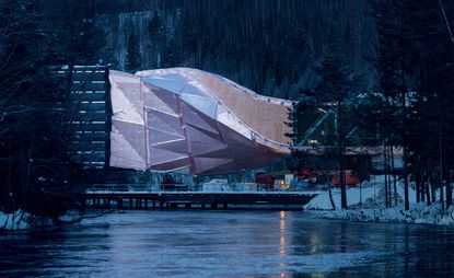 Exterior view of The Twist by Bjarke Ingels Group (BIG) during the construction phase. The twisted structure sits by a river and is surrounded by trees and snow