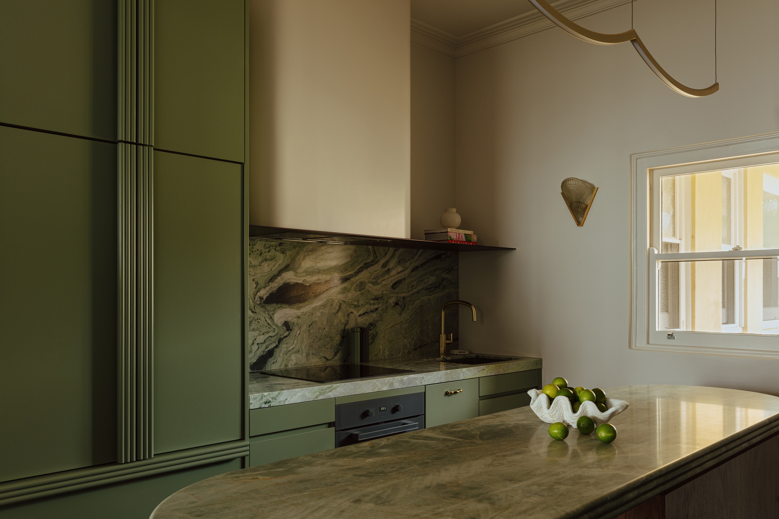 a small green kitchen with art deco inspired reeding on the cabinets, an art deco wall sconce, and a scallop bowl filled with limes