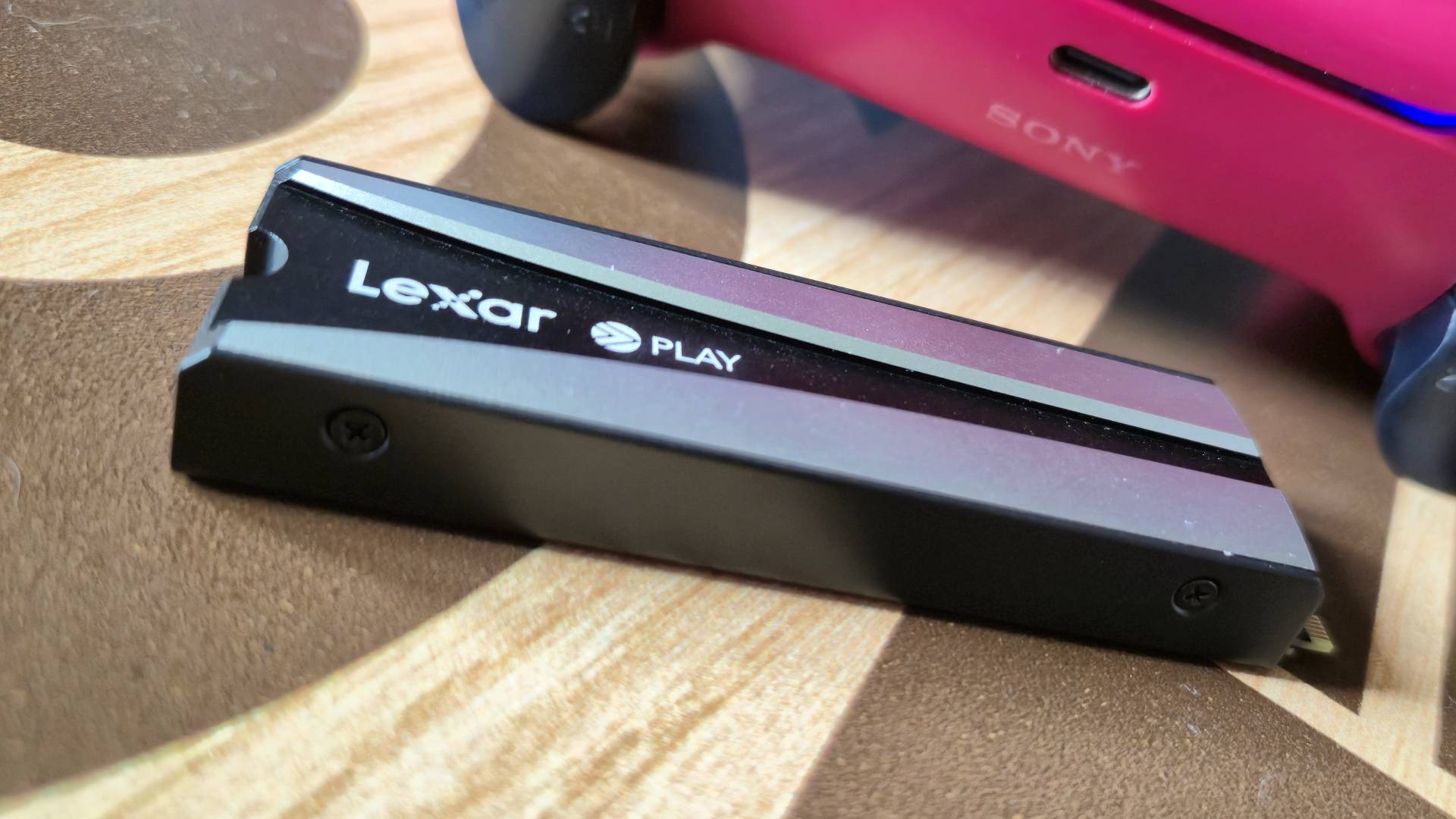 Photo by Rosalie Newcombe of the Lexar PLAY SSD sitting on a wooden table with a DualSense controller in the background to the right.