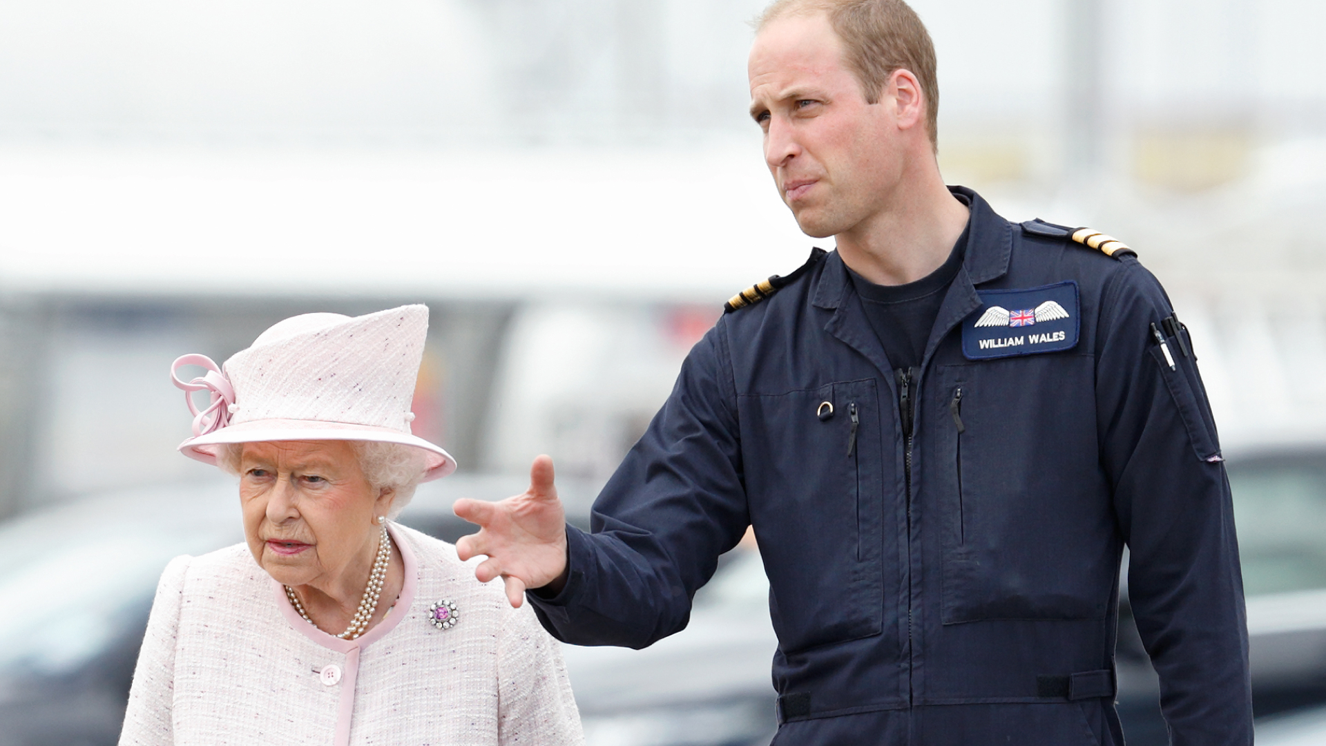 Queen Elizabeth wears a light pink coat and matching hat and carries a black leather handbag, while her grandson Prince William wears his Royal Air Force uniform and guides her around