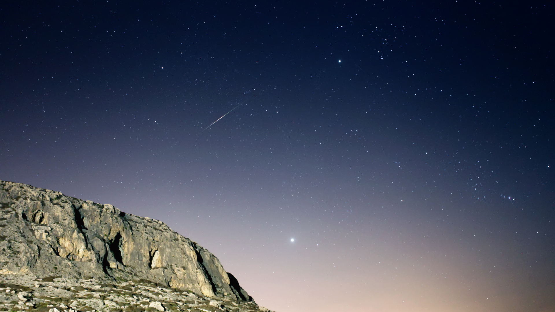 A purple and orange night sky filled with stars is seen behind a rocky cliff to the left of the image