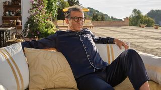 Jason Isaacs in blue pyjamas as Cary Grant looks pensive sitting on a yellow sofa on a patio in Archie.