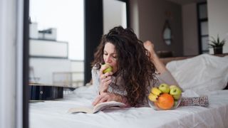 A woman lying on her front in bed next to a bowl of fruit, reading a book and eating the fruit