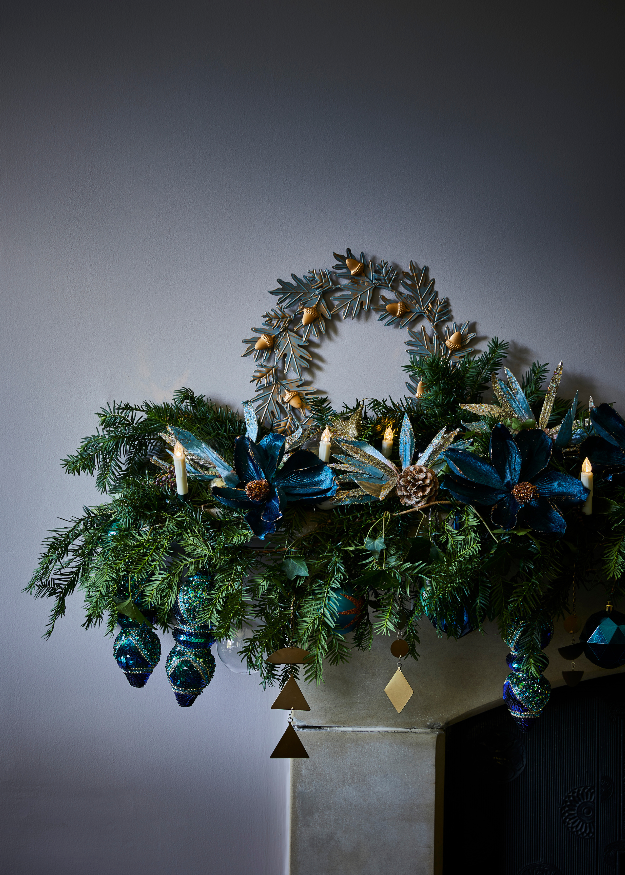 A christmas mantelpiece display covered in a garland with baubles decorated throughout and a metal wreath sitting behind all in a green blue and silver theme