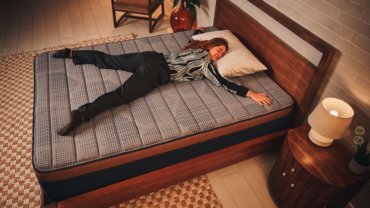 Our mattress tester in black and white patterned pyjamas laying on her front with arms and legs spread out on Brooklyn Bedding CopperFlex memory foam mattress on wooden bedframe with checkered rug and wooden bedside table with lamp