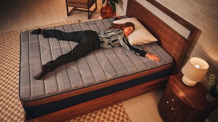 Our mattress tester in black and white patterned pyjamas laying on her front with arms and legs spread out on Brooklyn Bedding CopperFlex memory foam mattress on wooden bedframe with checkered rug and wooden bedside table with lamp