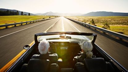 An older couple in a convertible car driving on a road in the sunshine