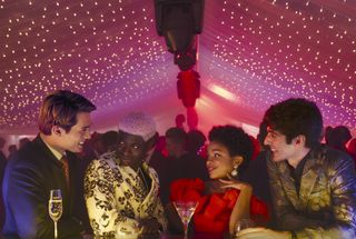 Henry (Nicholas Galitzine), Percy (Malcolm Atobrah), Nora (Rachel Hilson) and Alex (Taylor Zakhar Perez) celebrate New Year's Eve in a giant marquee, the ceiling filled with fairy lights. They are all smiling together, and there are glasses of champagne in front of them.