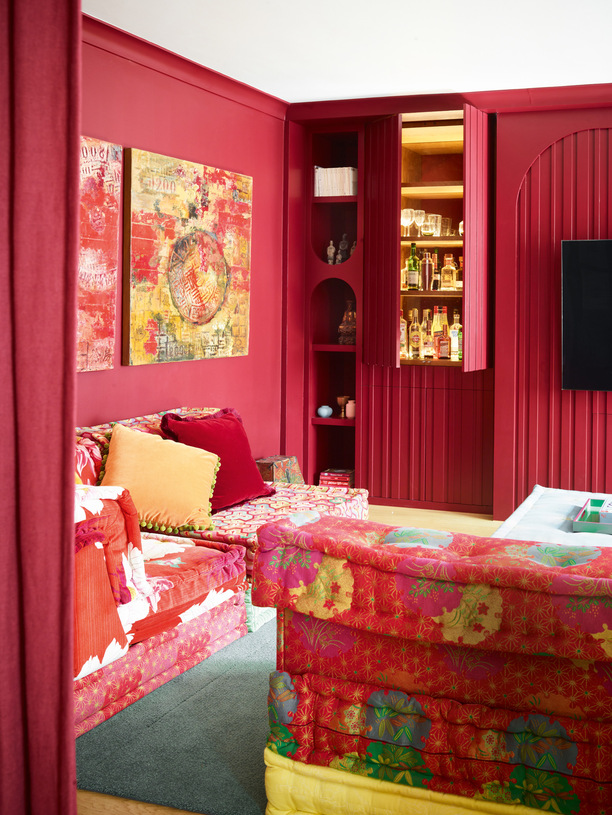 Deep pink-red living room with red, green and yellow floral patterned sofas, built-in bar with gold accents, and green rug