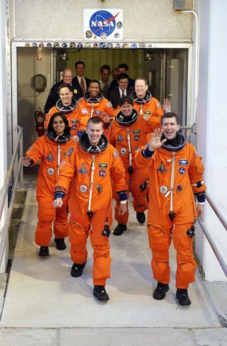 The astronauts are wearing bright orange flight suits and are smiling and waving.
