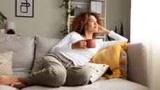 A young woman relaxes on her sofa with a cup of coffee and looks thoughtful.