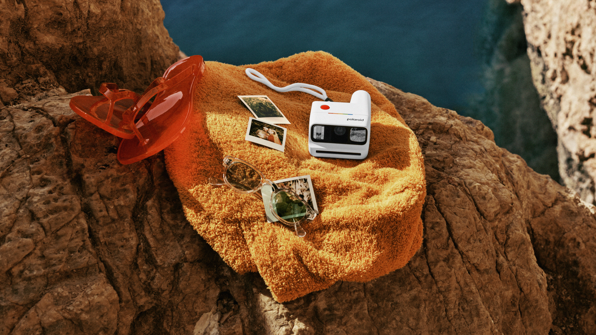 A Poalroid Go Gen 3 camera resting on a beach towel surrounded by instant prints and a pair of flip-flops, with the sea visible in the background