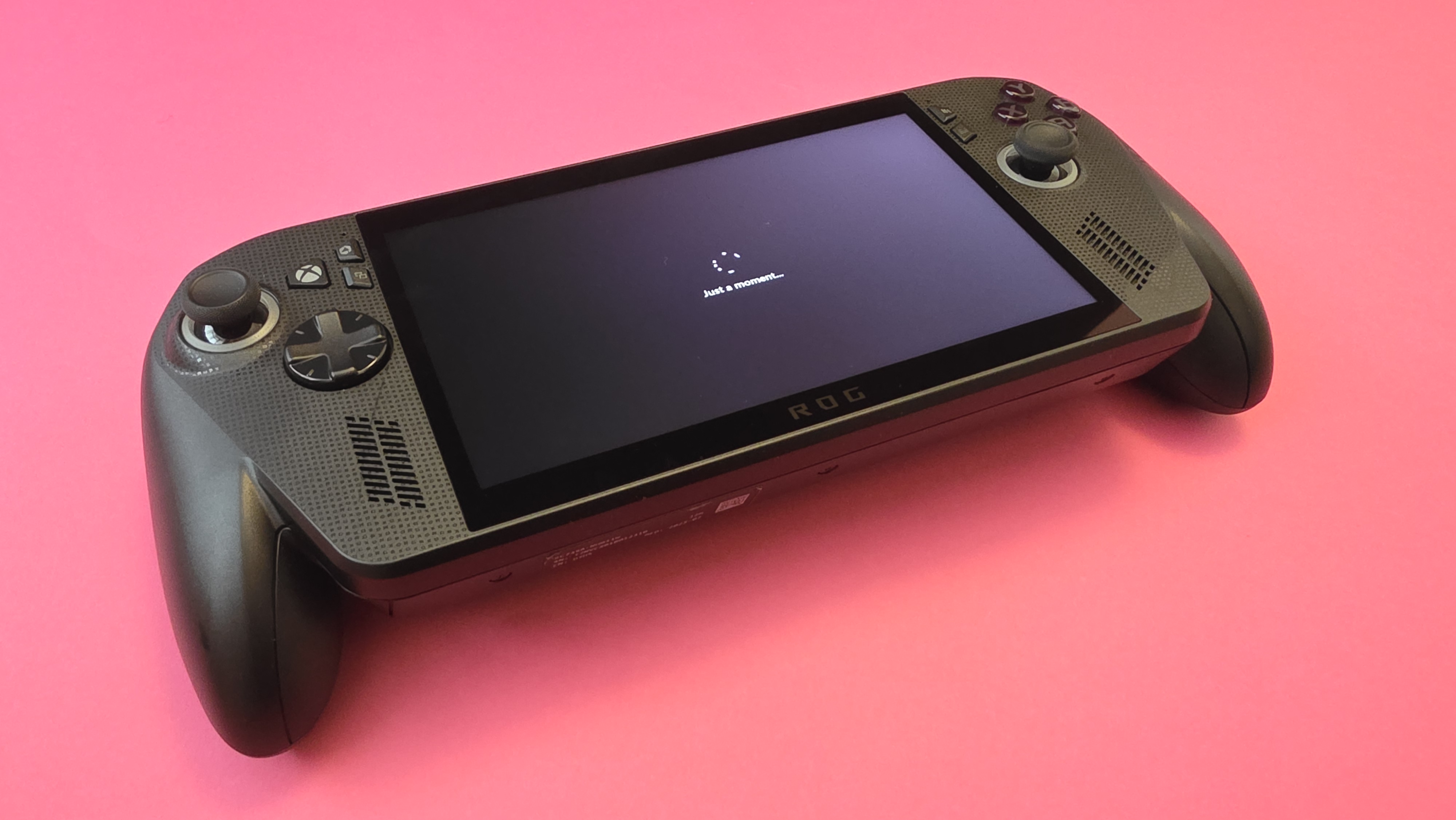 A handheld gaming device with a large screen and controls on each side rests on a pink surface.