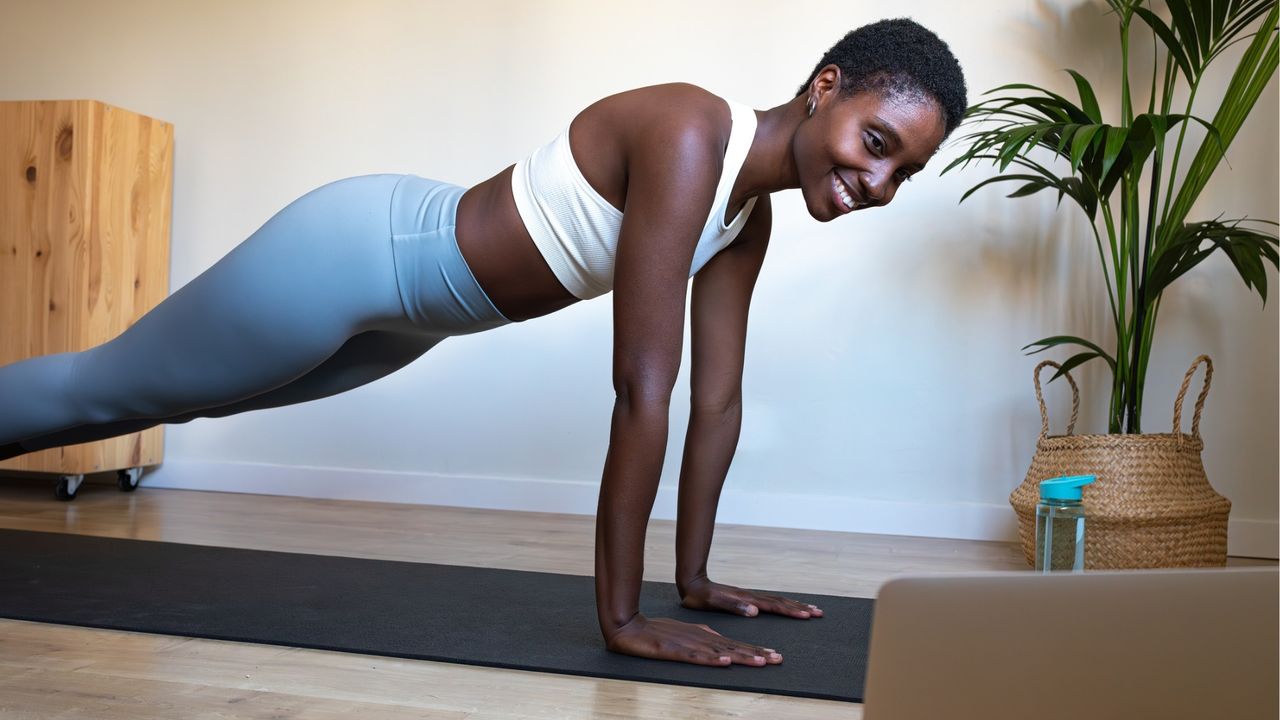 woman in a high plank position wearing white crop top and sky blue leggings smiling in front of laptop in room with plant in the corner 