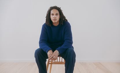 Portrait of Mac Collins sitting on chair with blue jumper and pale background