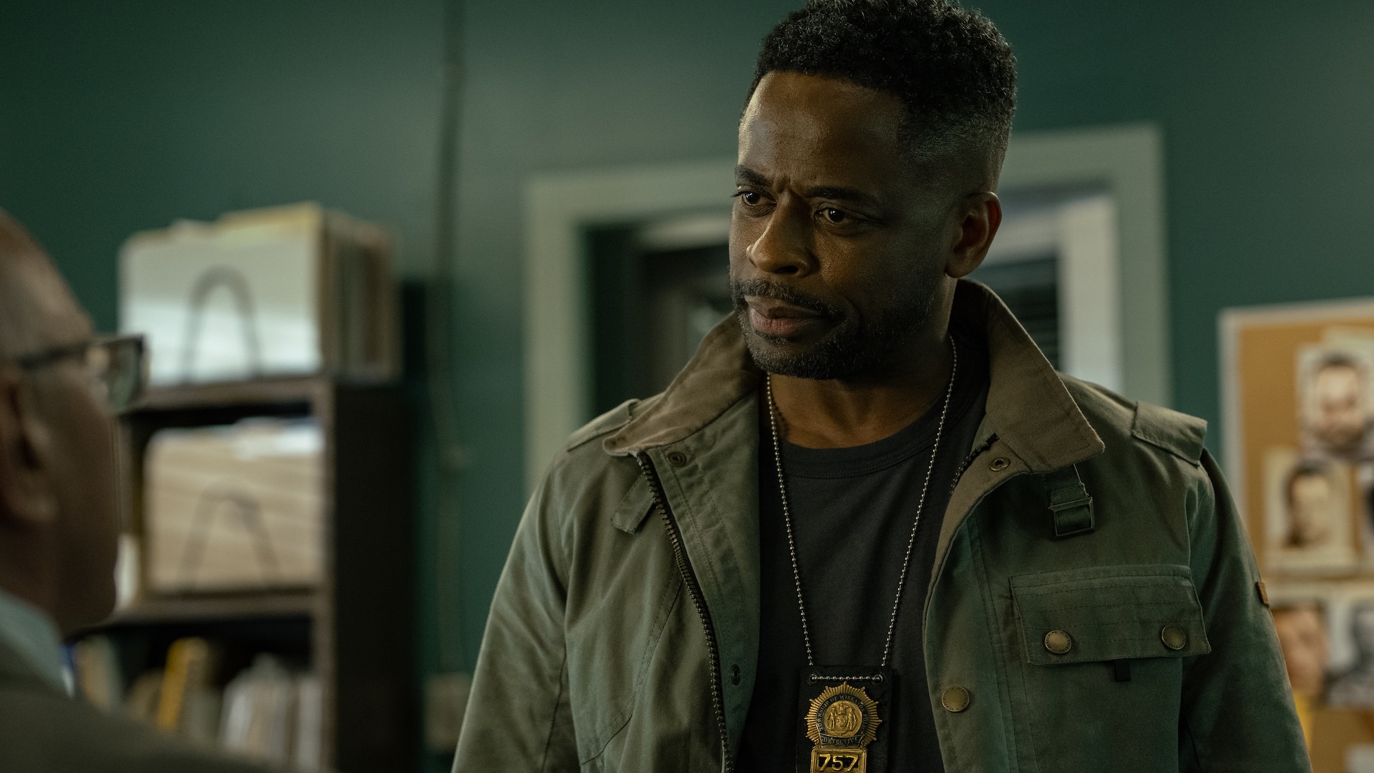 Dul&amp;eacute; Hill as Detective Rollins wearing green jacket and a badge hanging around his neck, standing in a police station with a bulletin board and file folders in the background in Yellowjackets Season 4