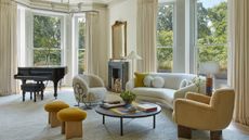 Image of a modern art nouveau living room. There are two large bay windows in the room with elegant, off-white curtains. There is a white curved sofa, a boucle white accent chair, a yellow accent chair, and two yellow stools. There is a round coffee table in the middle of the sitting area.