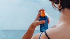 a wwoman's shoulder and the back of her head, pictured on a beach spraying on a blue bottle of sunscreen