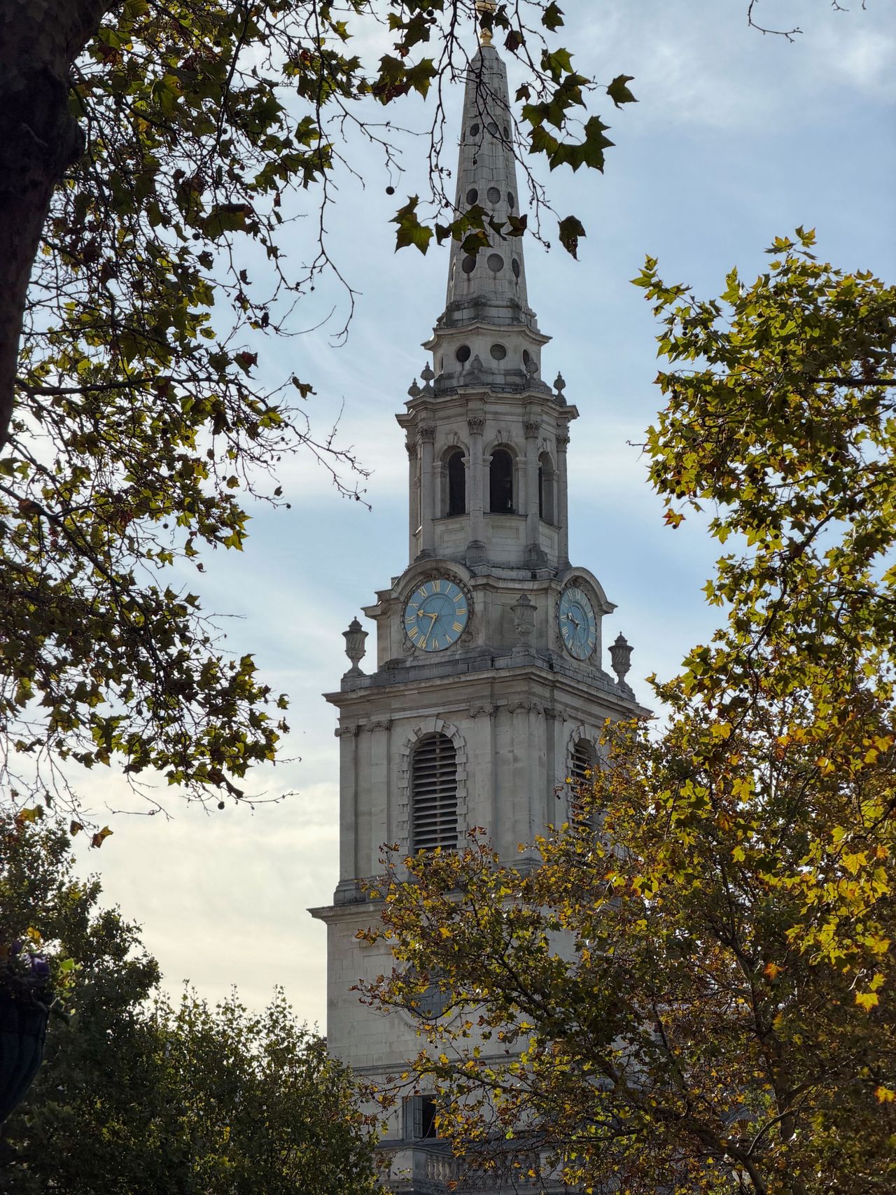 Ornate stone church clock tower and spire framed by autumn leaves against a pale blue sky.