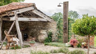 A rustic outdoor shower on green metro tiles beside a weathered wooden barn, gravel floor, Windsor chair, and lush potted plants.
