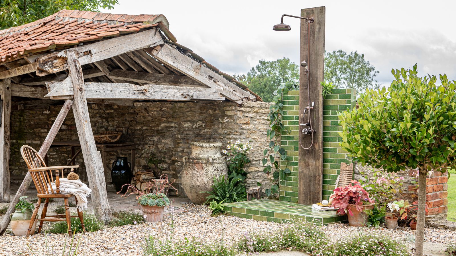 A rustic outdoor shower on green metro tiles beside a weathered wooden barn, gravel floor, Windsor chair, and lush potted plants.