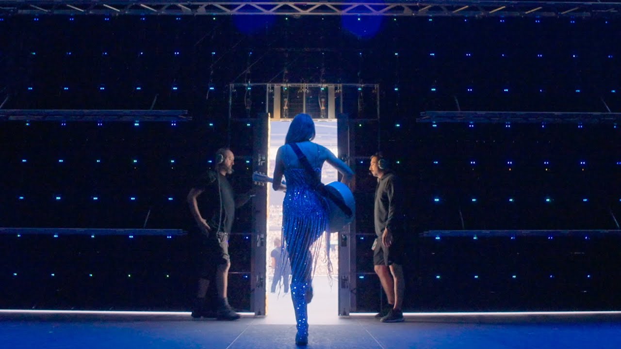 A shot from behind Taylor Swift, wearing a sparkly blue outfit and holding a guitar, as she walks onto a brightly lit stage between two crew members.