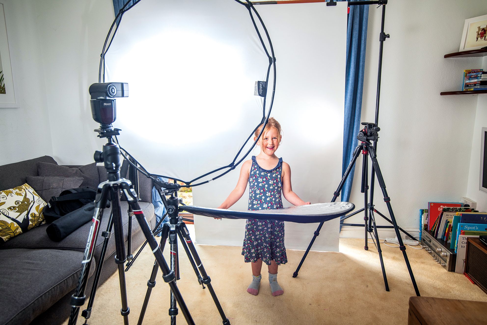 A child stands surrounded by photography equipment in a cozy room, holding a reflector while posing for a photo shoot